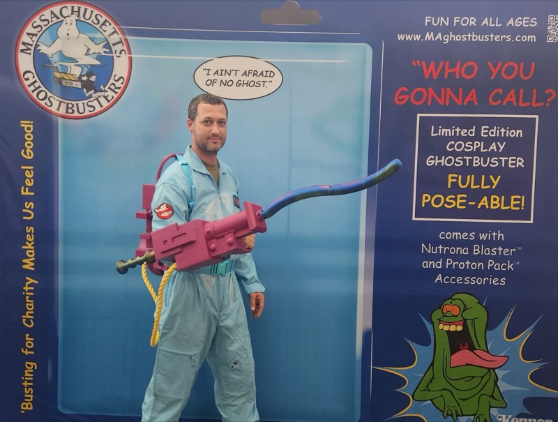 A convention participant holding a pink prop wand in front of the Massachusetts Ghostbusters toy-package backdrop.