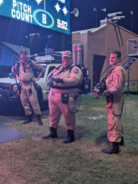 Three Ghostbusters fans posing with their gear beside a vehicle at a ballpark.