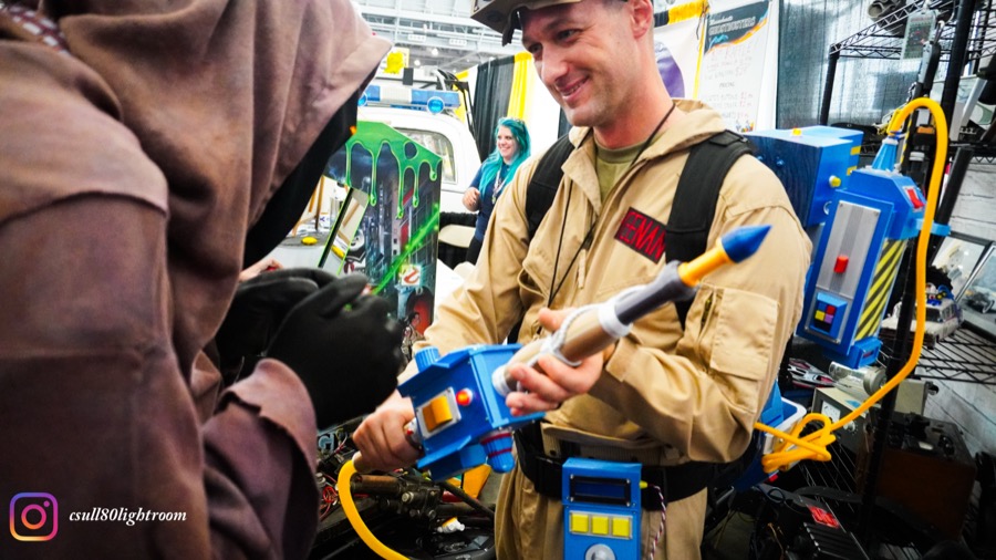 Convention attendee in a Ghostbusters uniform demonstrating blue-and-yellow prop gear at an event booth